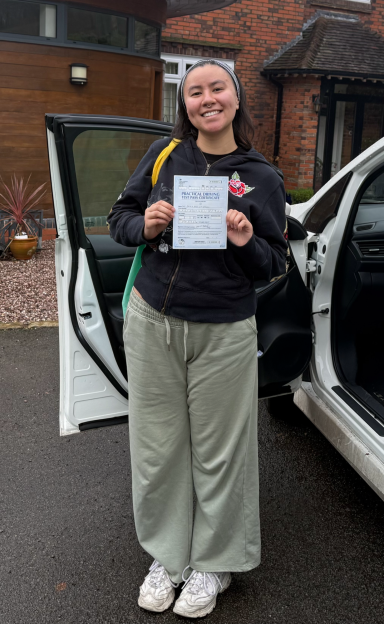Smiling person holding a paper outside a car, wearing casual clothes.
