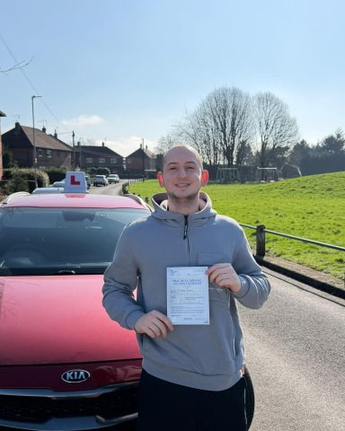 Person holding a certificate, standing in front of a red car on a sunny day.