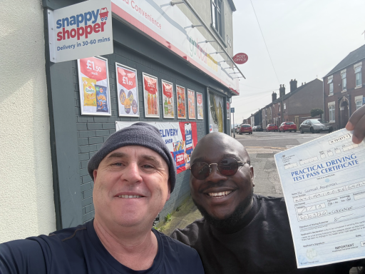 Two men smiling outside a shop, holding a certificate and posing for a selfie.