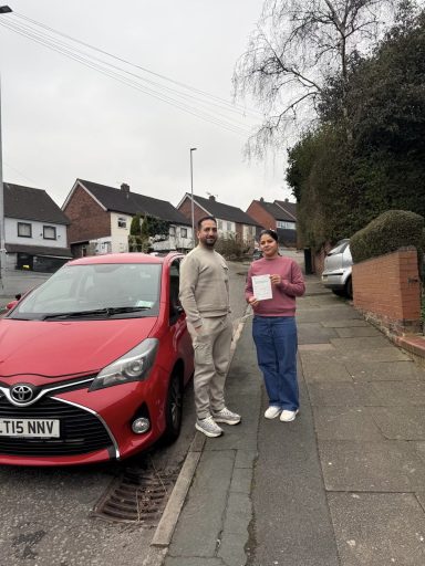 Two people stand next to a red car on a residential street, holding a document.