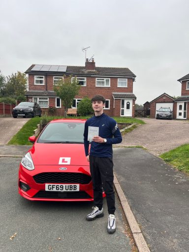 A person holding a driving test certificate stands next to a red car outside a house.
