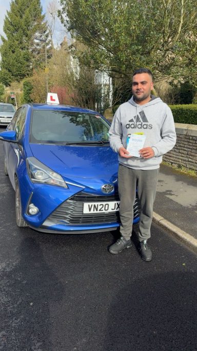 A person in a grey hoodie stands beside a blue car with a learner driver sign, holding a certificate.