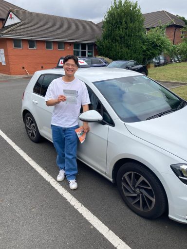 A young man stands by a white car holding a document, smiling outdoors.